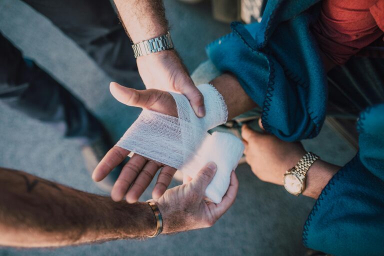 Cursus inhoud Close-up of two people bandaging an injured hand outdoors, focusing on first aid care.